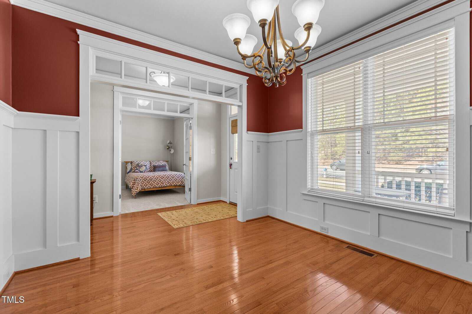 A vacant dining room with polished wooden floors, white wainscoting, and red upper walls. A brass chandelier hangs from the ceiling. Double glass doors open to a room with a bed. Large windows feature blinds, offering a view outside.