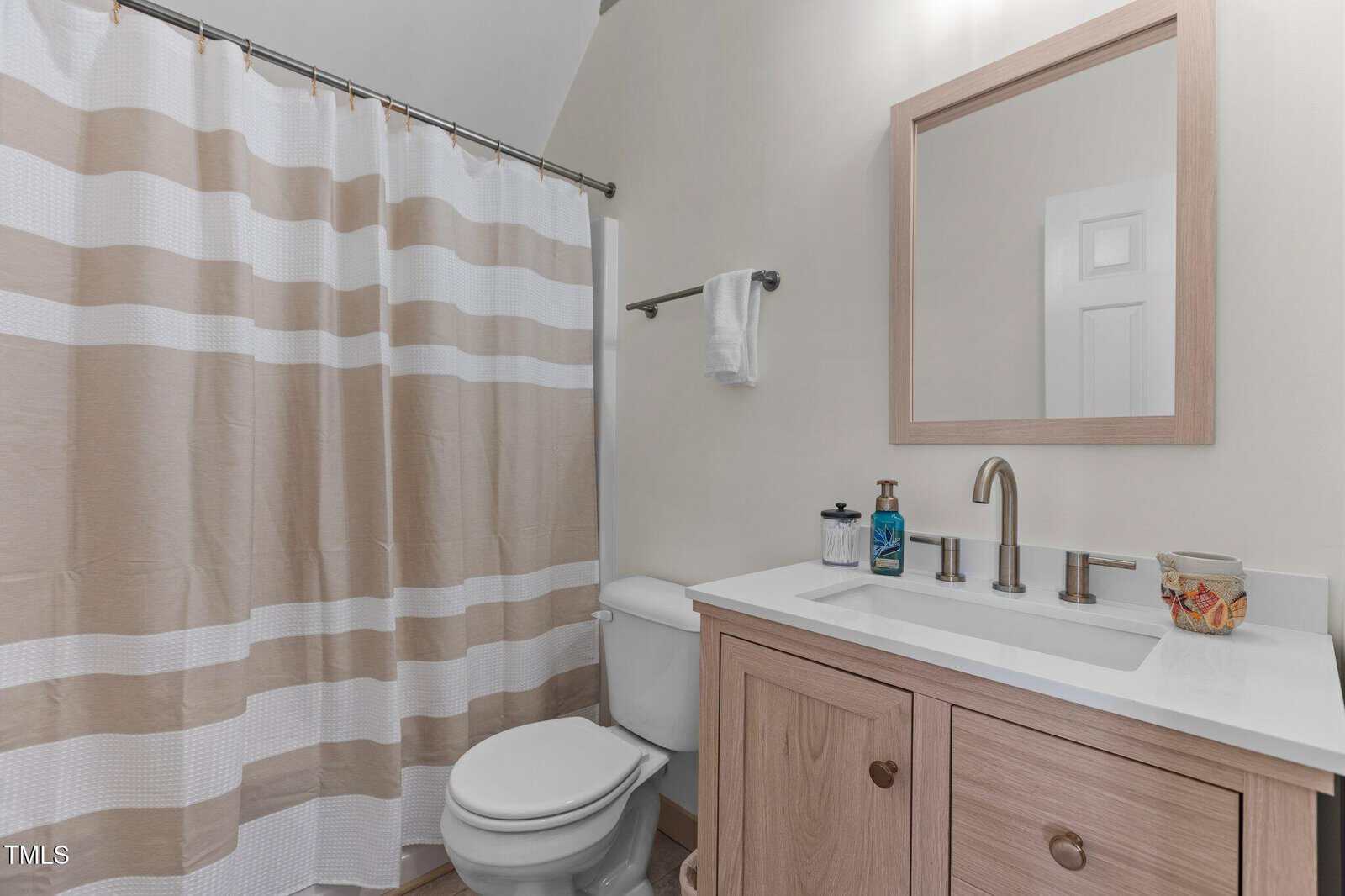Bathroom with a beige and white shower curtain, a white toilet, and a wooden vanity with a white countertop. A rectangular mirror is above the sink. A chrome faucet and soap dispenser are on the countertop, and a towel hangs on a rail.