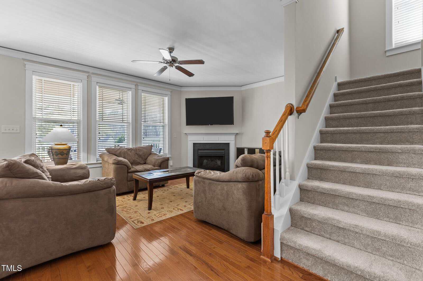 Living room with brown sofas, a wooden coffee table, and a carpet on hardwood flooring. There is a fireplace with a flat-screen TV above it, large windows with blinds, a ceiling fan, and a staircase with carpeted steps on the right.