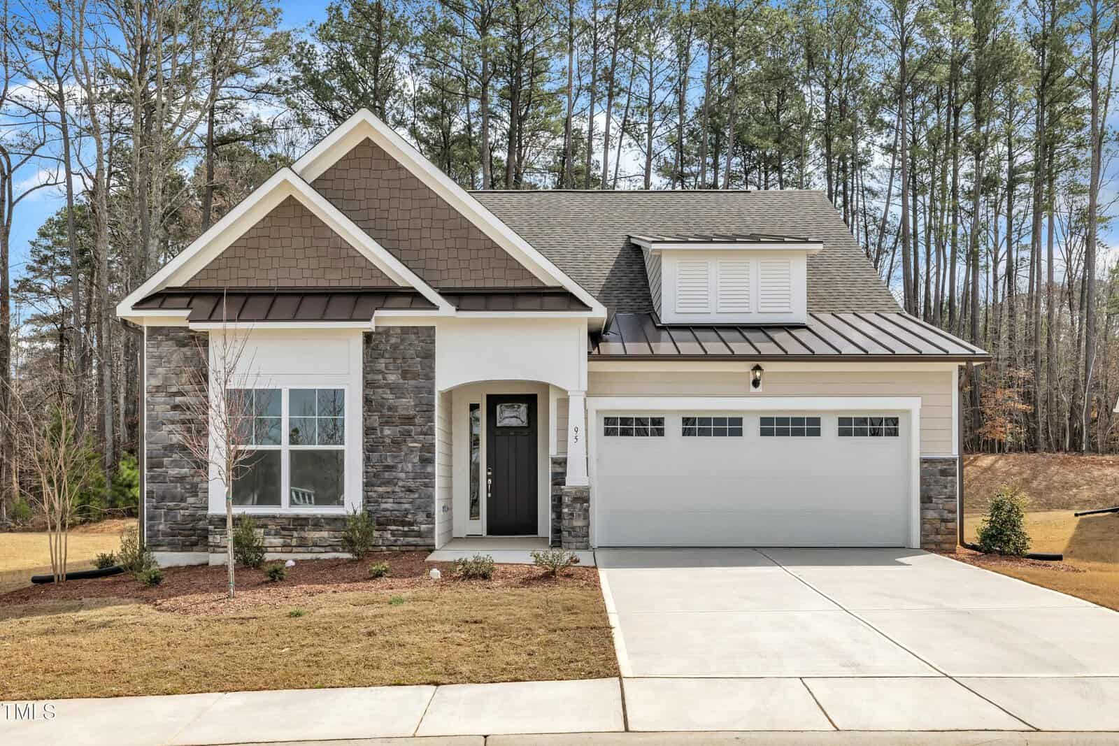 A modern single-family house with stone and siding exterior, covered porch, black front door, two-car garage, and a neatly landscaped yard. Tall trees are visible in the background under a partly cloudy sky.