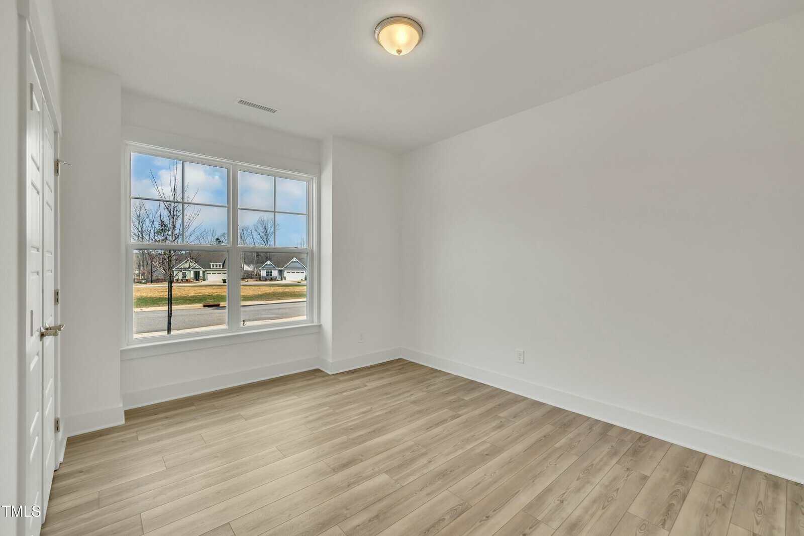 Empty room with light-colored wood flooring, white walls, a ceiling light fixture, and a large window letting in natural light. The window offers a view of houses and trees outside.