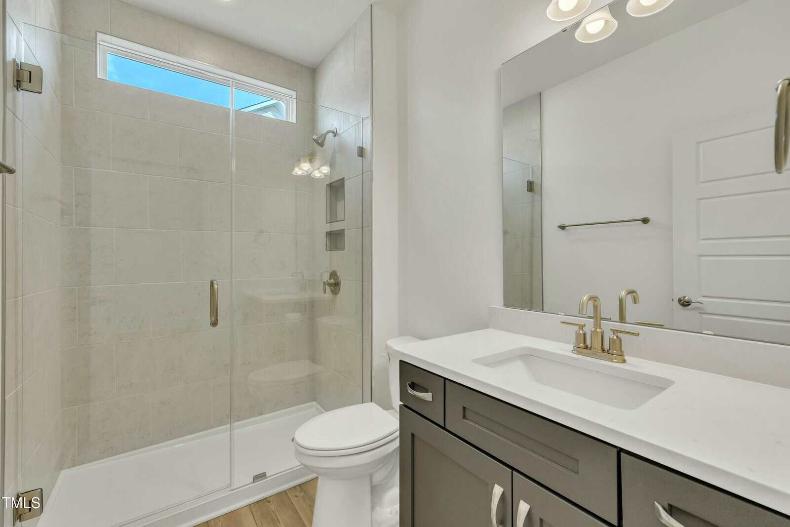 Modern bathroom with a glass-enclosed shower, white toilet, dark vanity with white countertop, large mirror, gold fixtures, towel rack, and a high window above the shower letting in natural light.