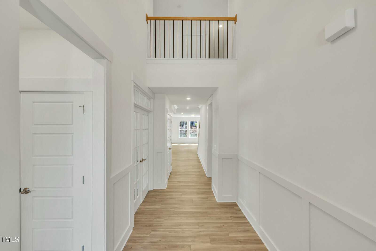A hallway with light-colored walls and wood flooring, featuring white doors on the left, wainscoting, a railing above, and a view into a bright room with large windows at the end.