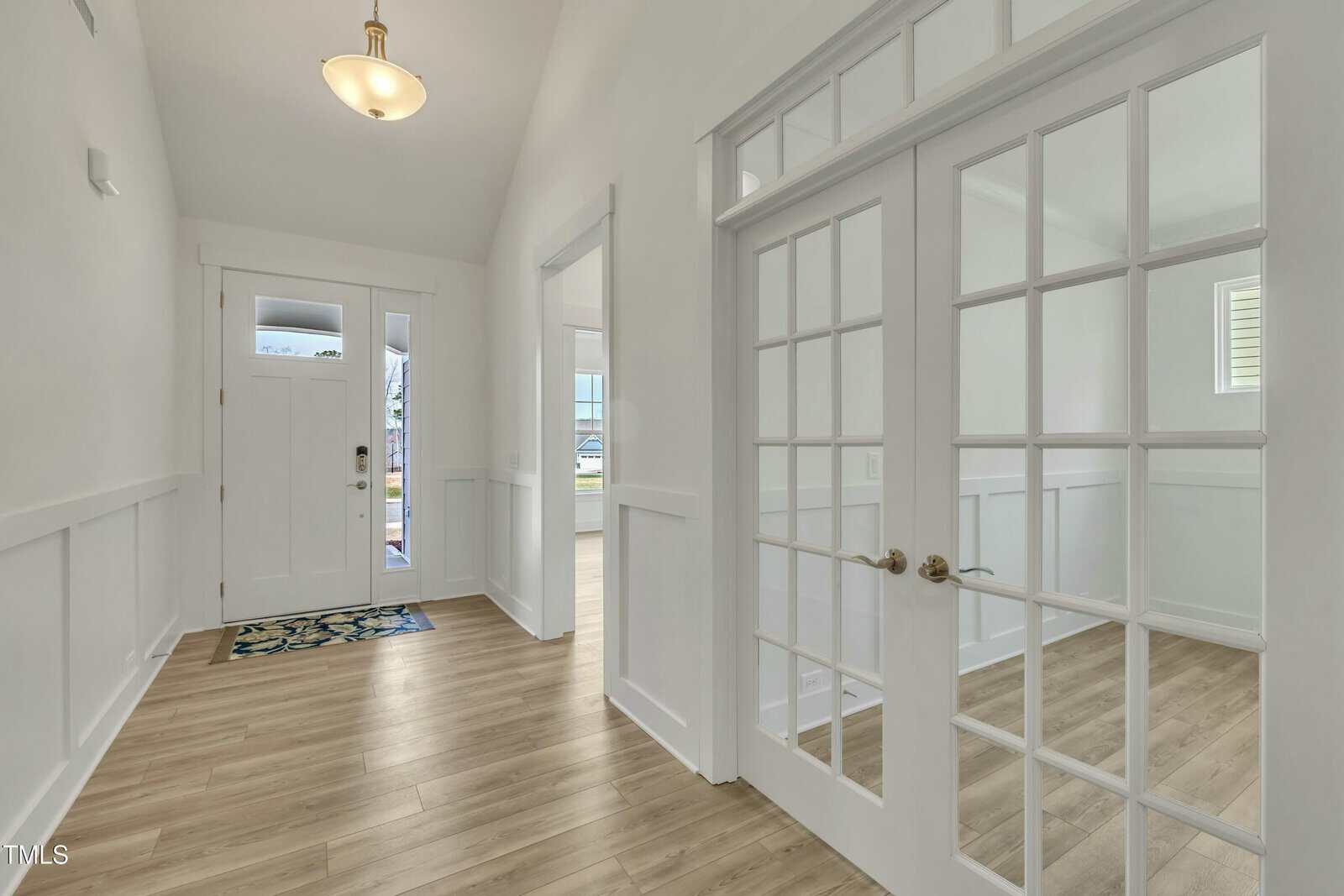 Bright entryway with light wood flooring, white walls, a front door with sidelights, and double French doors leading into an adjacent room; a ceiling light fixture hangs above, and a patterned rug is near the door.