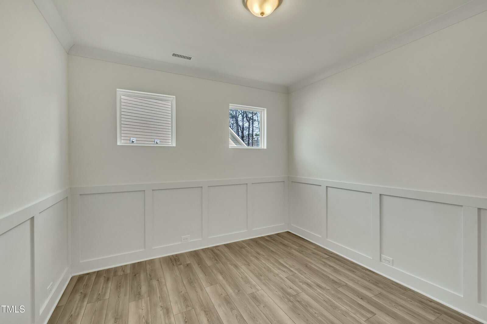 Empty room with light-colored walls, white trim, wood-style flooring, two small rectangular windows, and a ceiling light fixture. The room has wainscoting and a simple, modern appearance.