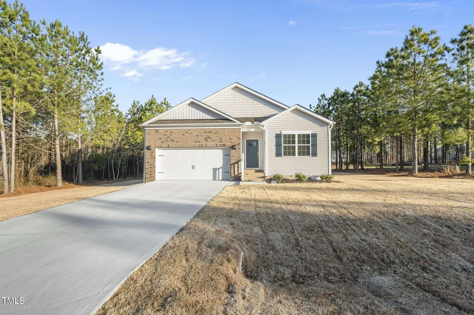A single-story house with a brick and white exterior, attached two-car garage, concrete driveway, and a lawn, surrounded by pine trees under a clear blue sky.