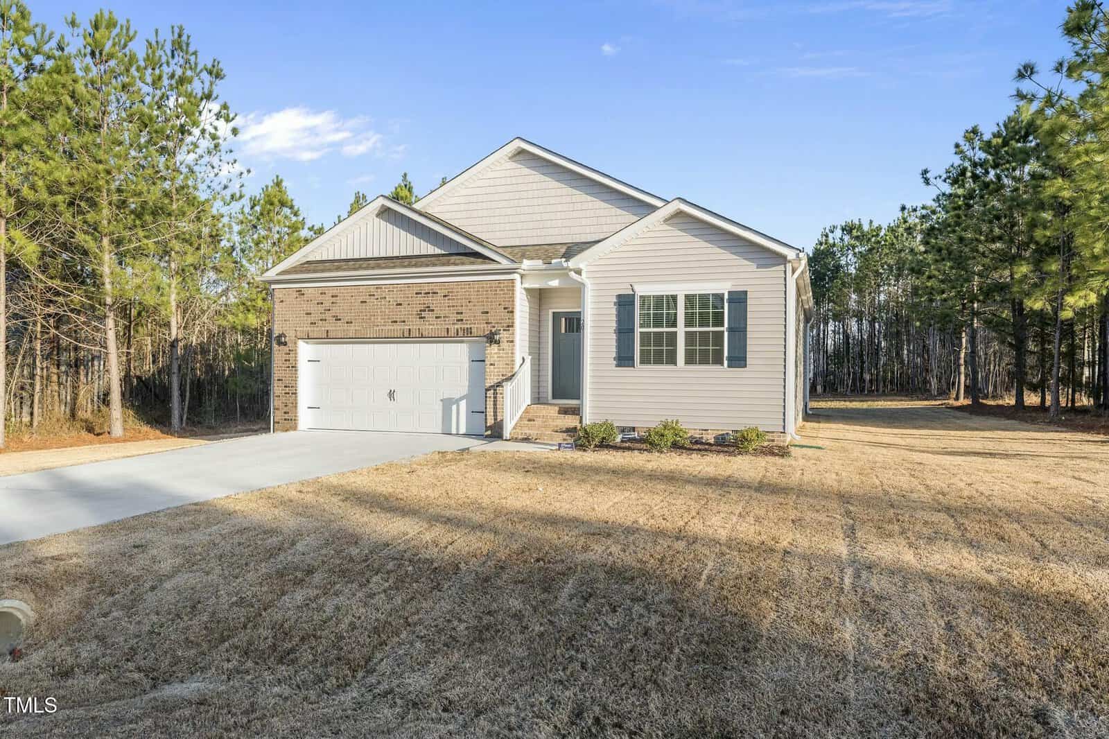 Single-story house with a brick and siding exterior, two-car garage, and small front porch. The house is surrounded by a large yard with grass and bordered by pine trees under a clear blue sky.
