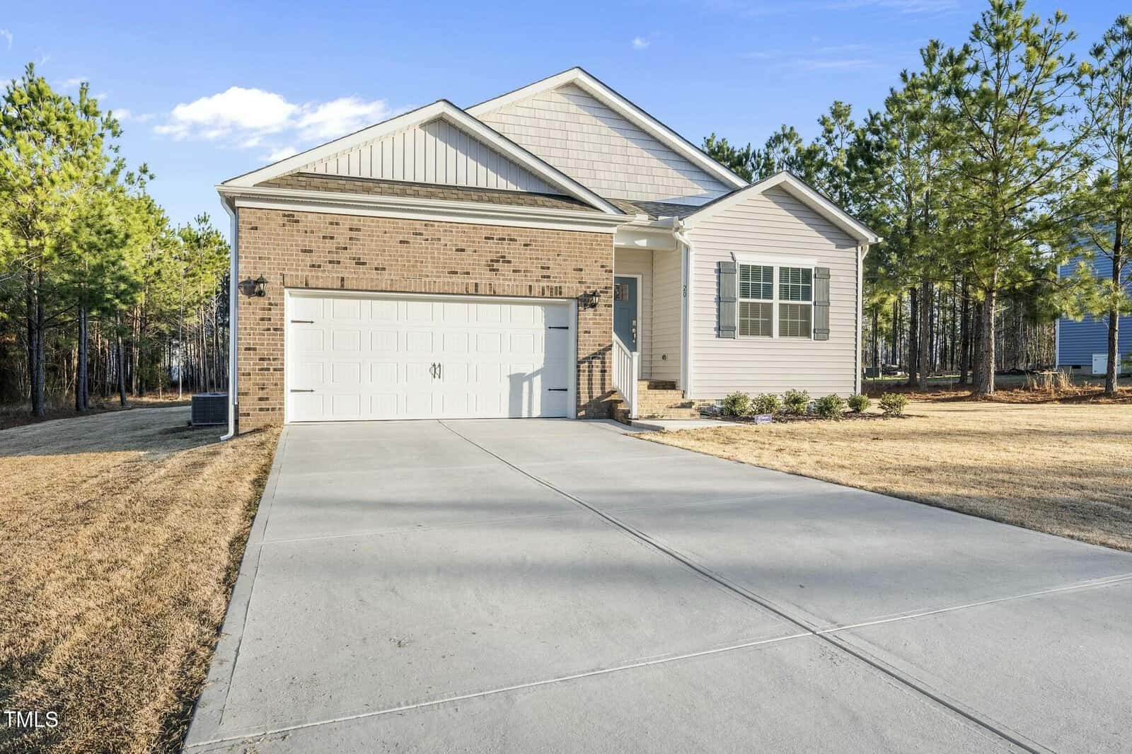 A single-story house with a brick and siding exterior, a two-car garage, and a wide concrete driveway, surrounded by pine trees and dry grass under a clear blue sky.