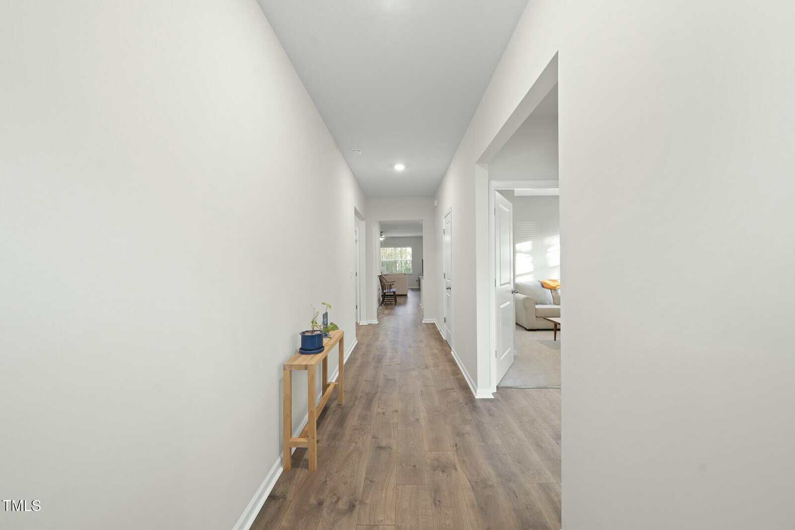 A long, bright hallway with light-colored walls and wood flooring. A simple wooden table with a few plants is on the left, and an open doorway on the right leads to a living room with a sofa.