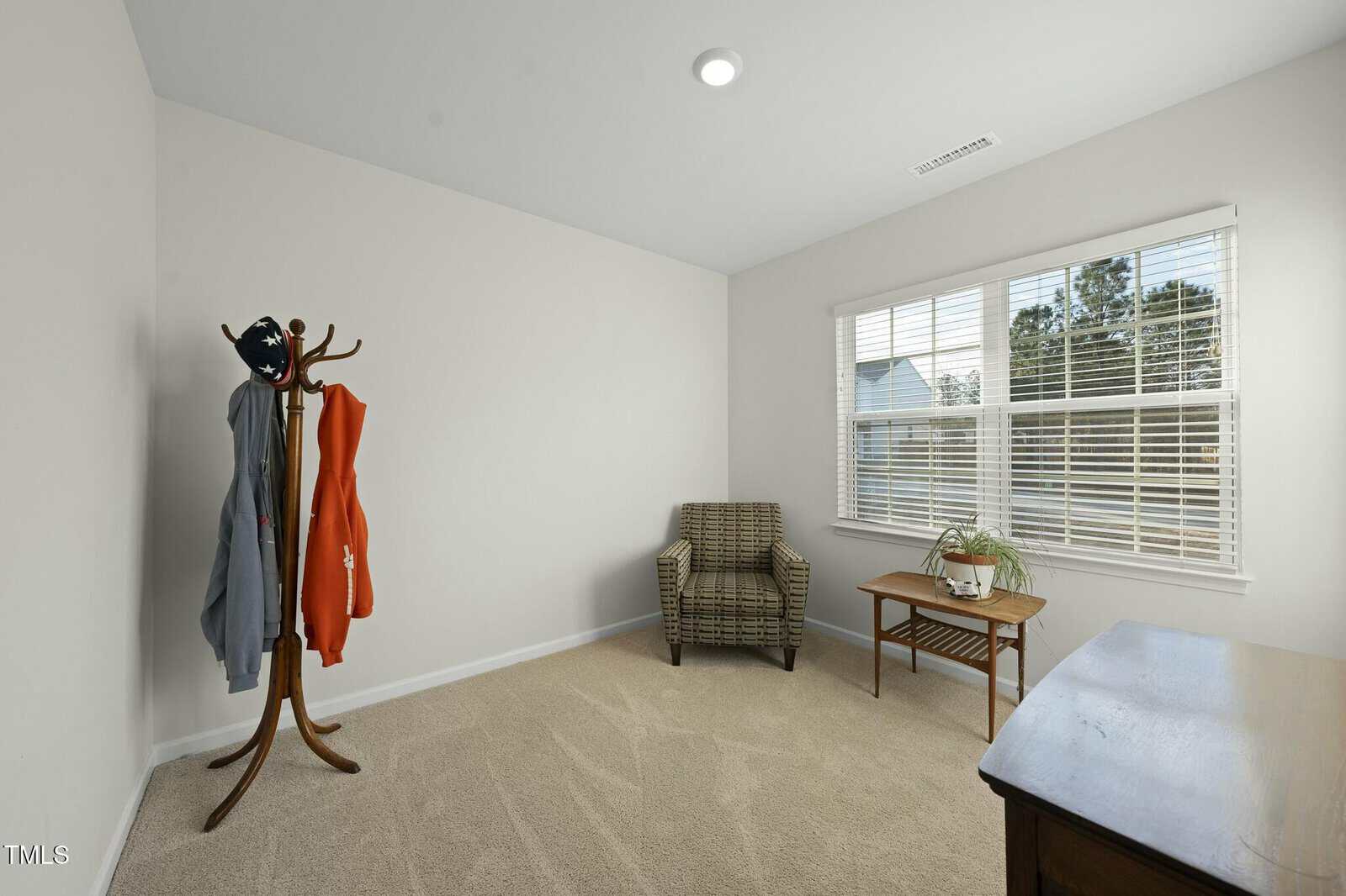 A small, carpeted room with white walls features a checkered armchair, a wooden coat rack with clothes, a side table with a plant, and a wooden desk. Sunlight enters through a window with white blinds.