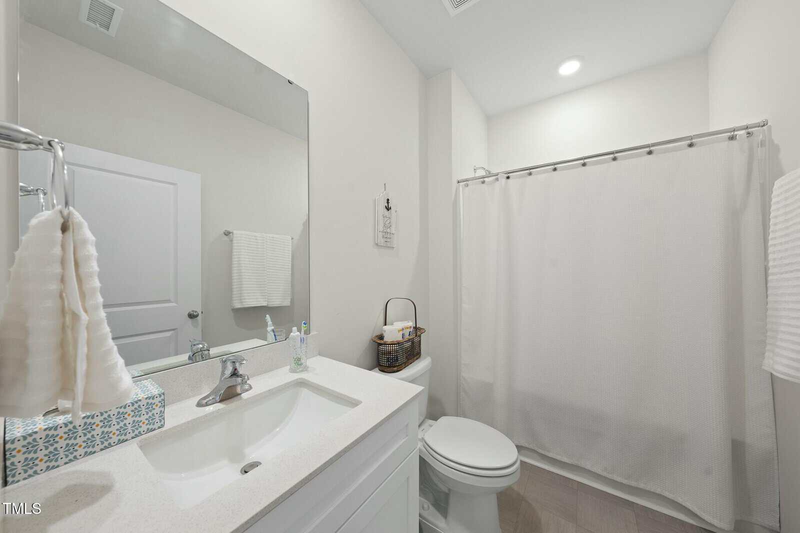 A modern bathroom with a white sink, silver faucet, tissue box, soap dispenser, and towel on the left. There is a toilet, basket with supplies, and a bathtub with a white shower curtain in the background.
