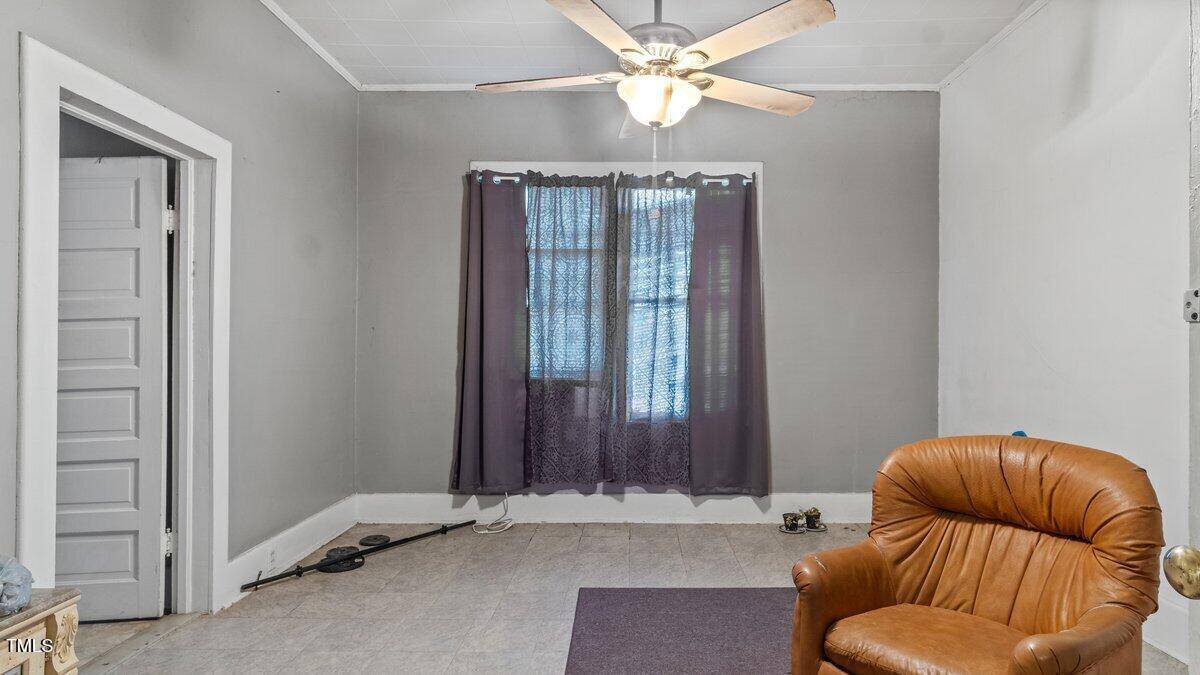 A small room with gray walls and a white ceiling fan. There is an armchair in the foreground, and a window with sheer black curtains in the background. A partially open door is on the left, and there are wires on the floor.