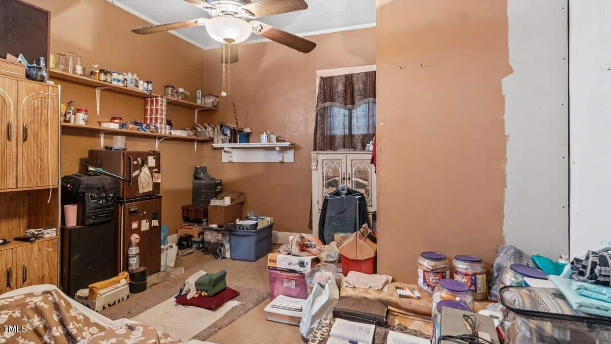 A cluttered room with brown walls and a ceiling fan. Various items are scattered, including bins, bags, and storage containers. Shelves with jars and cans line one wall. A small brown fridge and a wooden dresser are also present.