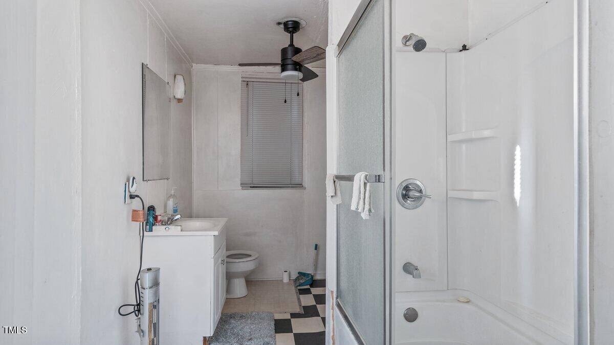 A compact bathroom with a square black and white checkered floor. It features a glass-enclosed shower, a sink with a mirror above, a toilet, a ceiling fan, and a small window with blinds. Some toiletries are visible on the sink.