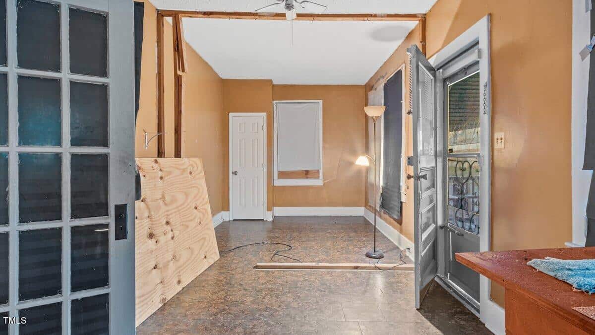 An empty room with tan walls and a white ceiling features a floor lamp, an open doorway leading outside, a covered window, and a closed door. There is a wooden board propped against the wall and a long plank on the floor.