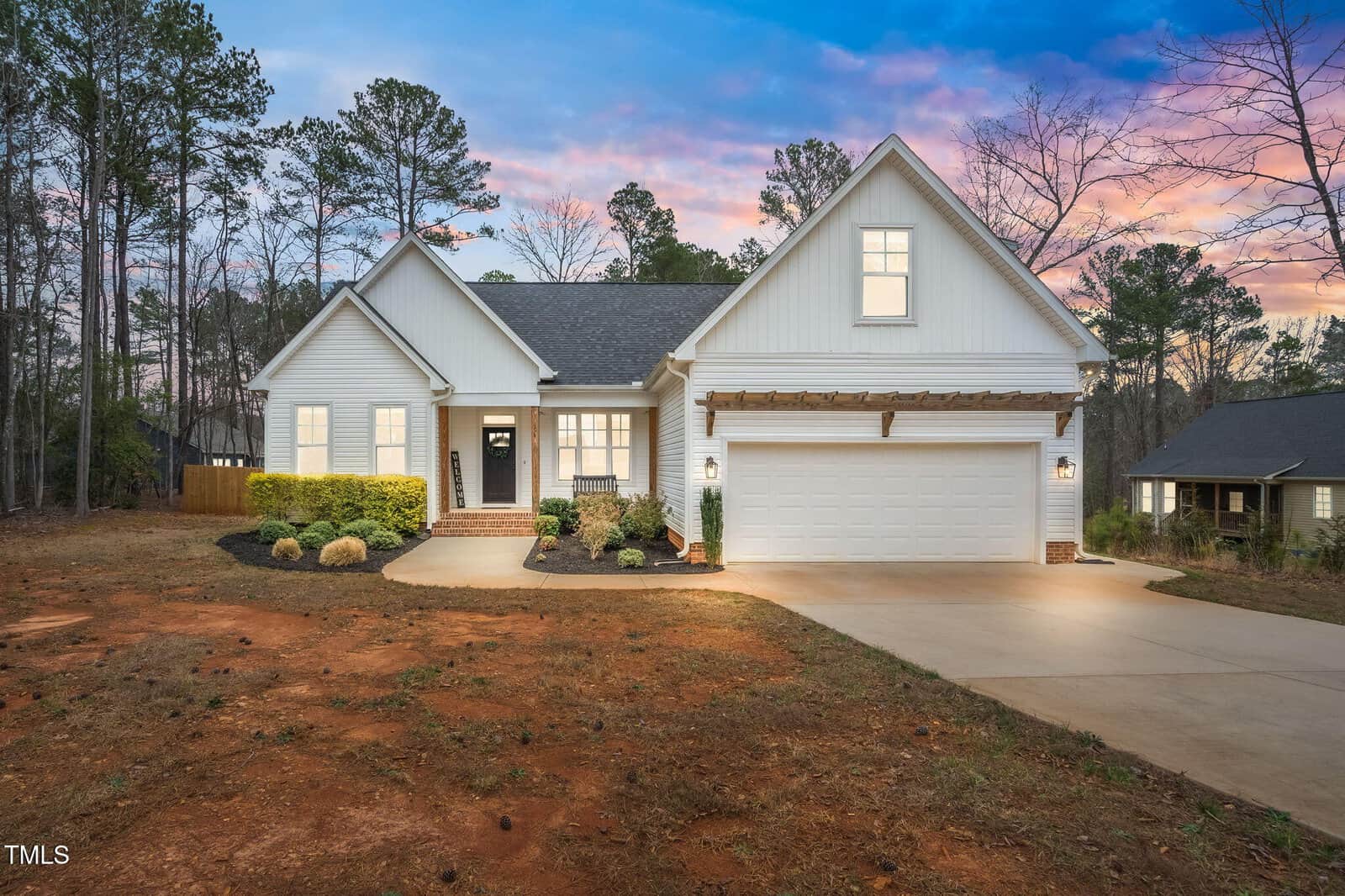 A white single-story house with a two-car garage, front porch, and landscaped yard sits on a concrete driveway. Tall trees and a neighboring house are visible in the background during sunset.