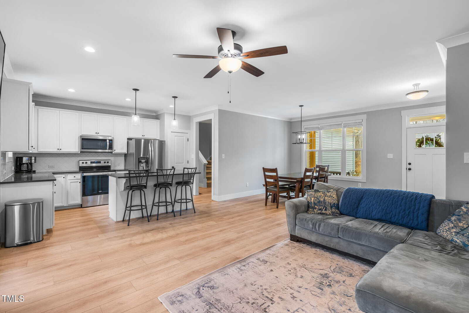 Open-concept living area with a gray sofa, dining table, and kitchen featuring white cabinets, stainless steel appliances, and a center island with barstools. Ceiling fans and wood floors are present throughout the space.