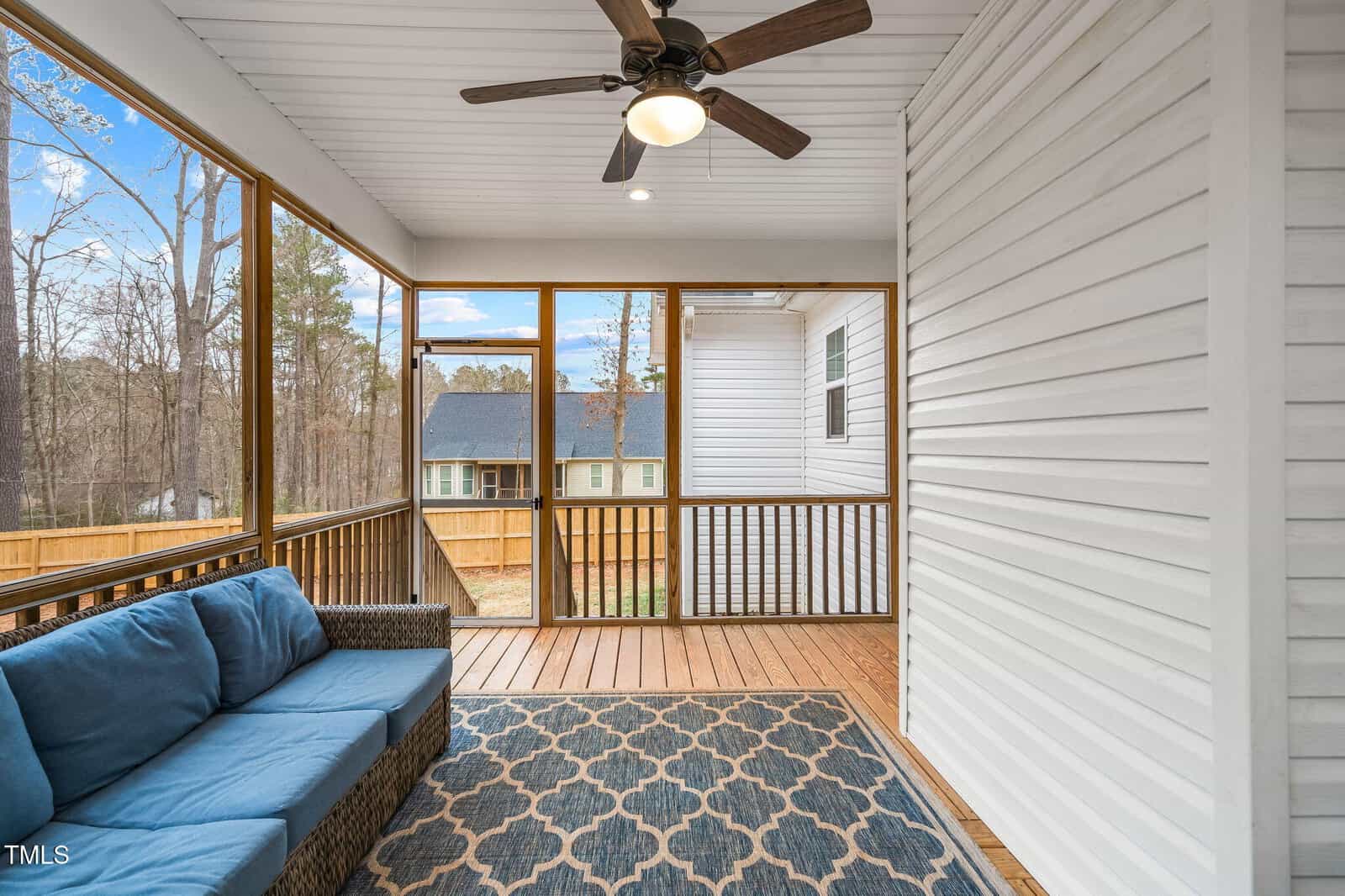 A screened-in porch with a ceiling fan, wood floor, patterned rug, wicker sofa with blue cushions, and white siding walls. The porch overlooks a backyard with a wooden fence and trees in the background.