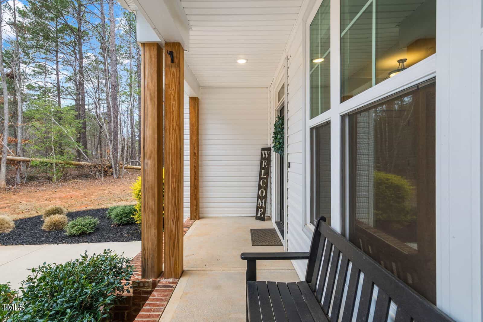 A covered front porch with a black bench, a “Welcome” sign by the door, wooden pillars, and landscaped plants. Large windows reflect the interior lighting. Trees and shrubs are visible in the background.