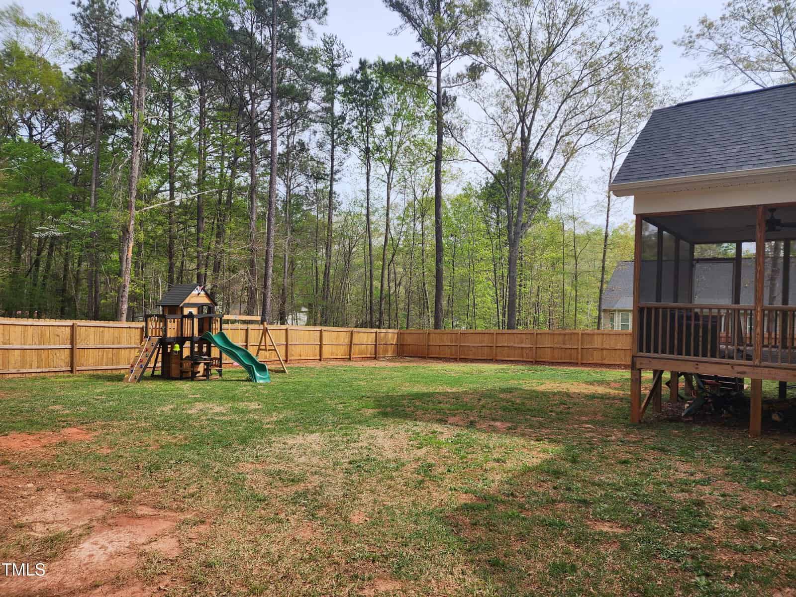 A fenced backyard with a patchy lawn, a wooden playset with a slide and swings on the left, and a screened porch attached to a house on the right. Tall trees are visible beyond the fence.