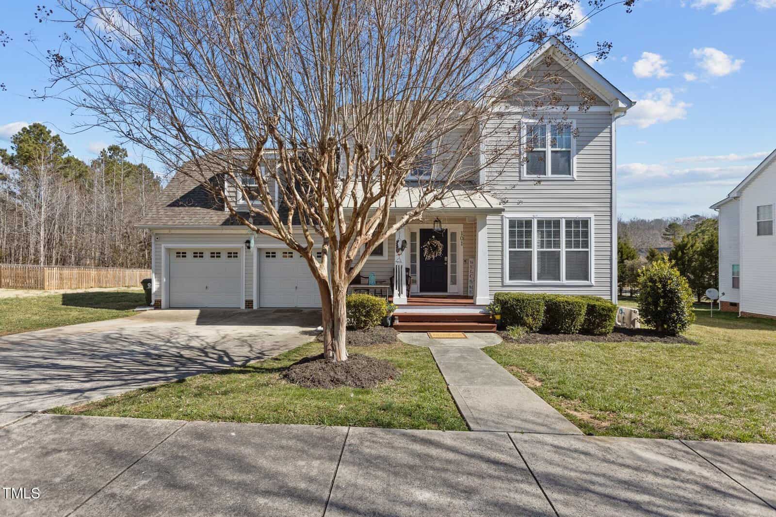 Front view of a two-story house at 101 Cambridge Way, Oxford, showcasing gray siding and white trim. The property features a two-car garage and paved driveway. A leafless tree graces the porch area, surrounded by well-maintained shrubs and grass under a clear, sunny sky.