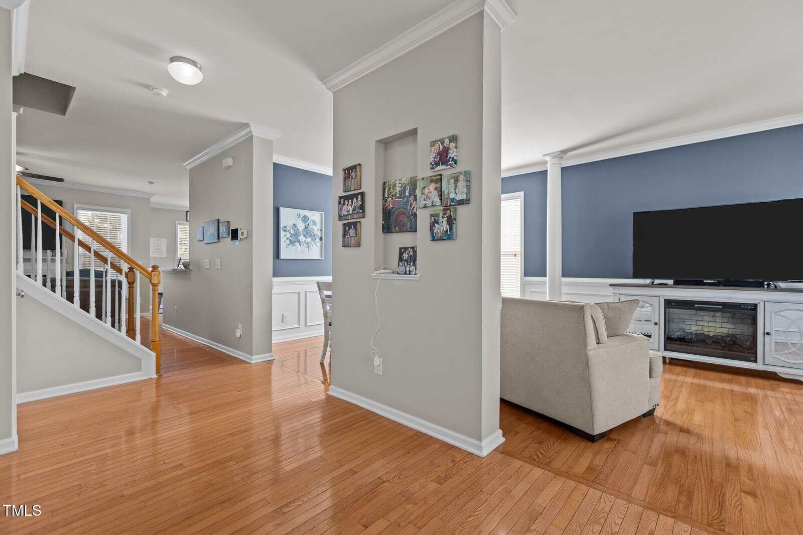 Interior of a home with a hallway and living room. The hallway features a wooden staircase and photo collage on the wall. The living room has blue walls, a large TV, a light-colored couch, and a fireplace. Wood flooring is throughout the area.