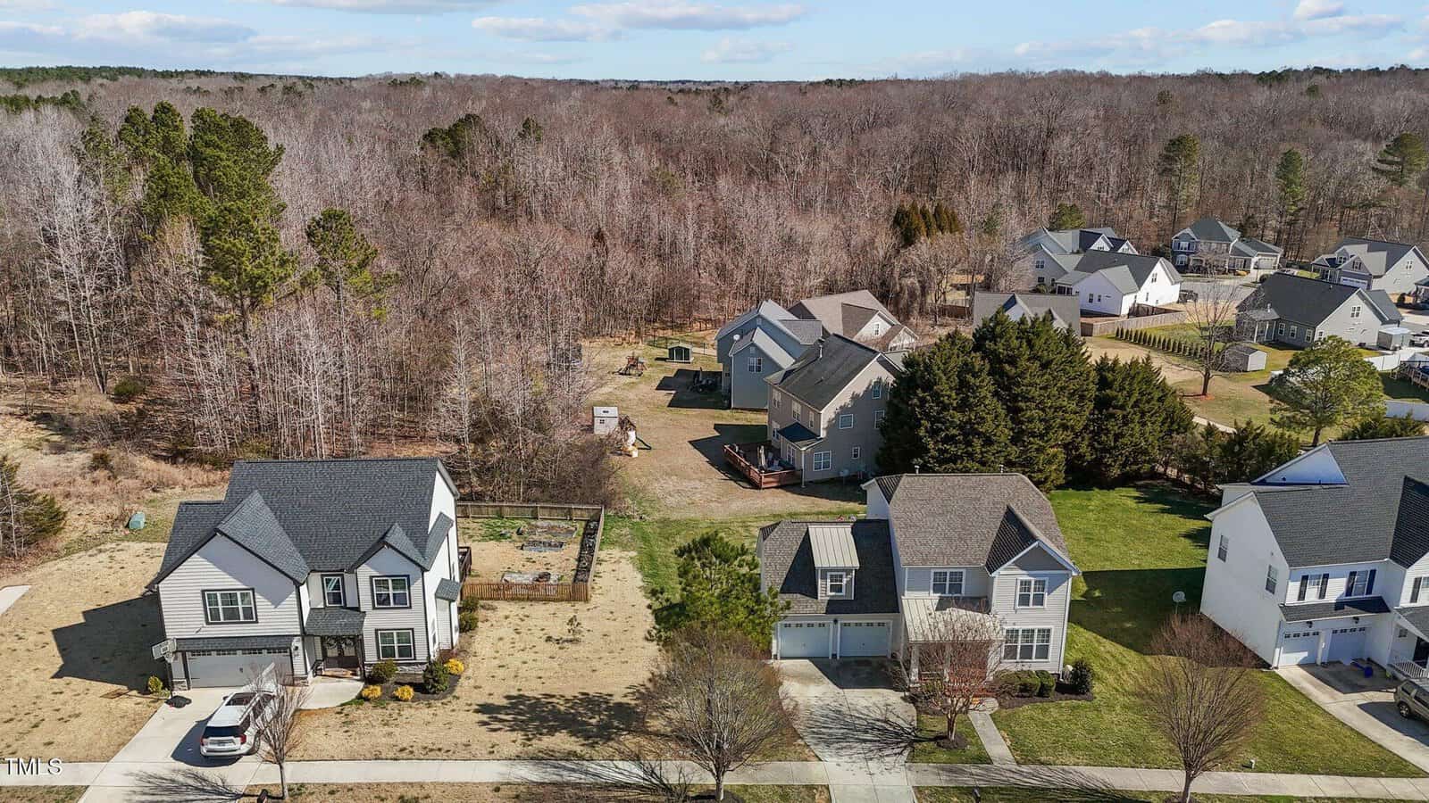 Aerial view of a suburban neighborhood featuring several houses with driveways and yards. Some homes have fences and decks. The background shows a wooded area with bare trees under a partly cloudy sky.