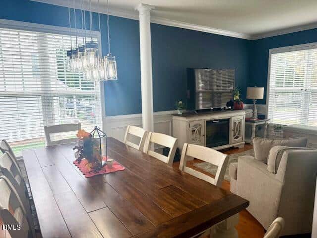 A dining area with a wooden table and eight white chairs is next to a living space with a sofa, TV, and console. Blue walls, large windows with blinds, and a modern glass light fixture are visible.