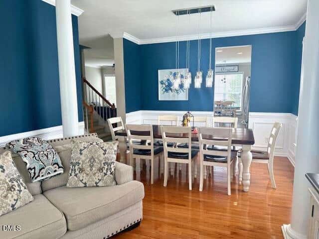 A dining room with blue walls, white wainscoting, and a rectangular wooden table with eight chairs. A beige sofa with patterned pillows is in the foreground. Pendant lights hang above the table.