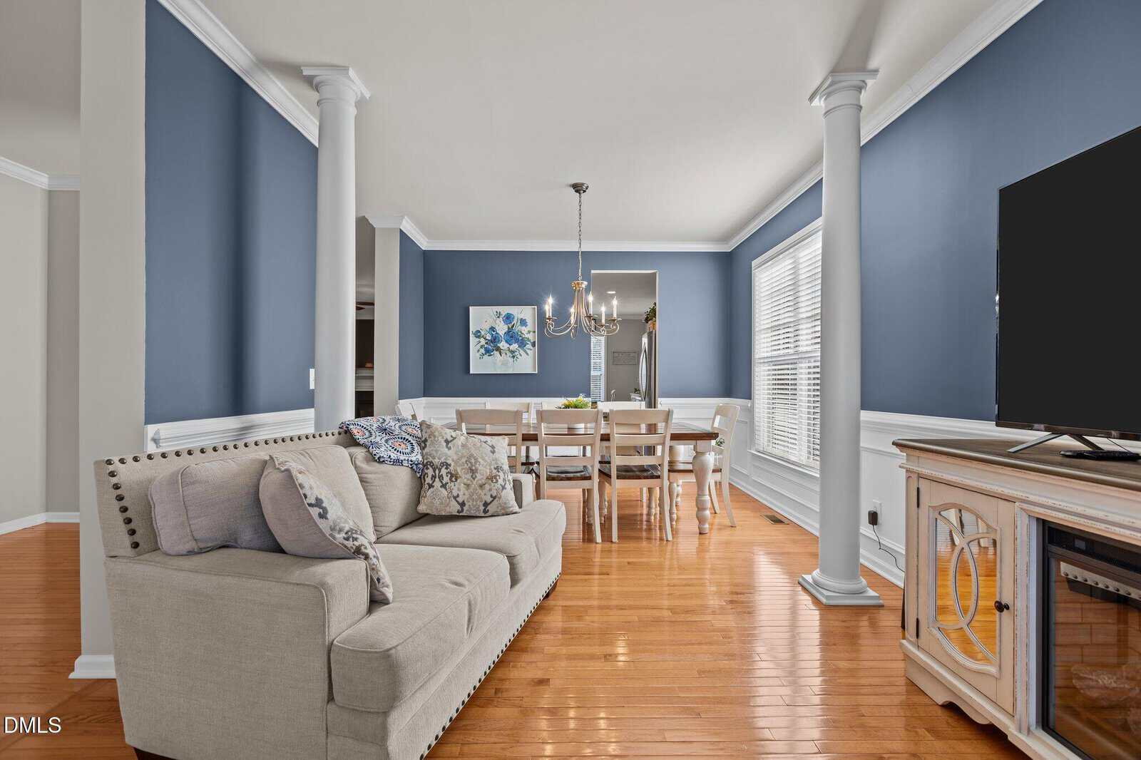 A living and dining room with blue walls, white trim, wooden floors, a beige sofa with patterned pillows, a dining table with six chairs, a chandelier, and a TV on a console. Large windows let in natural light.