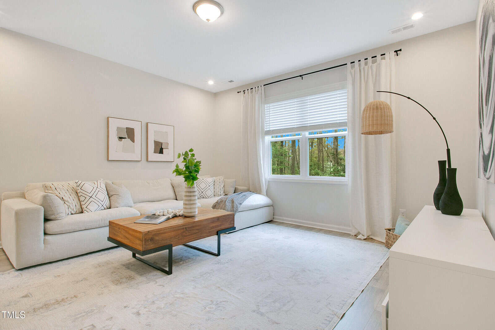 A bright living room with a white sectional sofa, a wooden coffee table, wall art, a large window with white curtains, a floor lamp, and a white rug covering most of the floor.