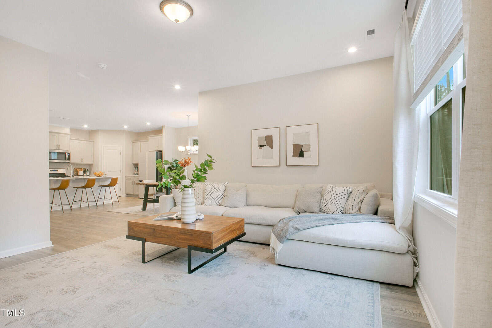 A modern living room with a light sectional sofa, wooden coffee table, abstract wall art, and large windows. The space opens to a kitchen and dining area with barstools and contemporary decor.