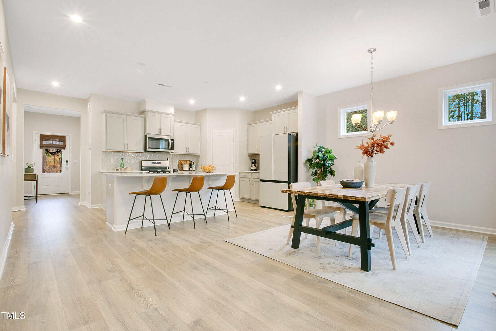 A modern, open-concept kitchen and dining area with light wood floors, white cabinets, a kitchen island with three barstools, a dining table with chairs, and large windows letting in natural light.