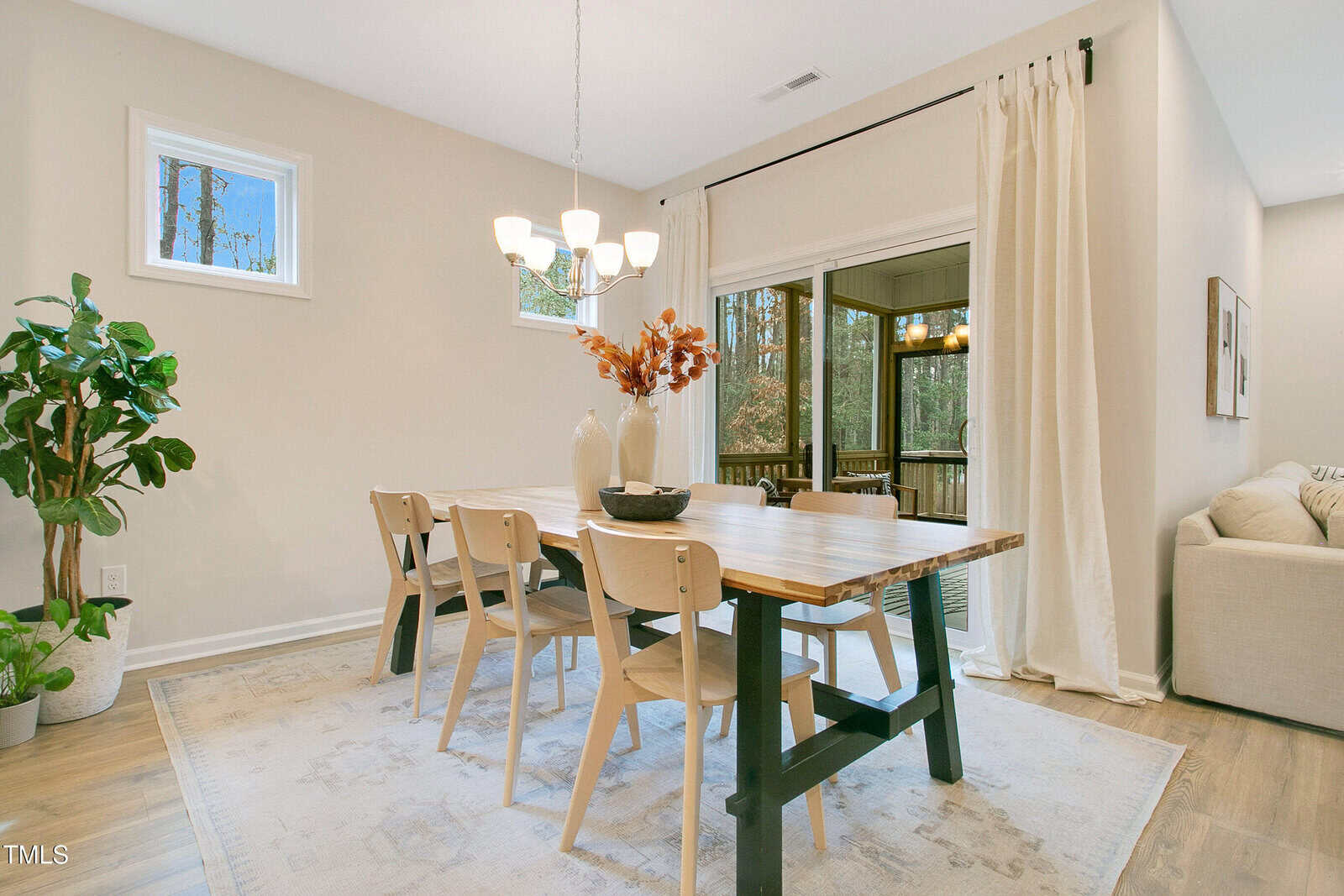 A dining room with a wooden table, six light wood chairs, a white rug, two vases with dried orange leaves, a bowl centerpiece, a chandelier, a large window with curtains, a plant, and an adjacent screened porch.