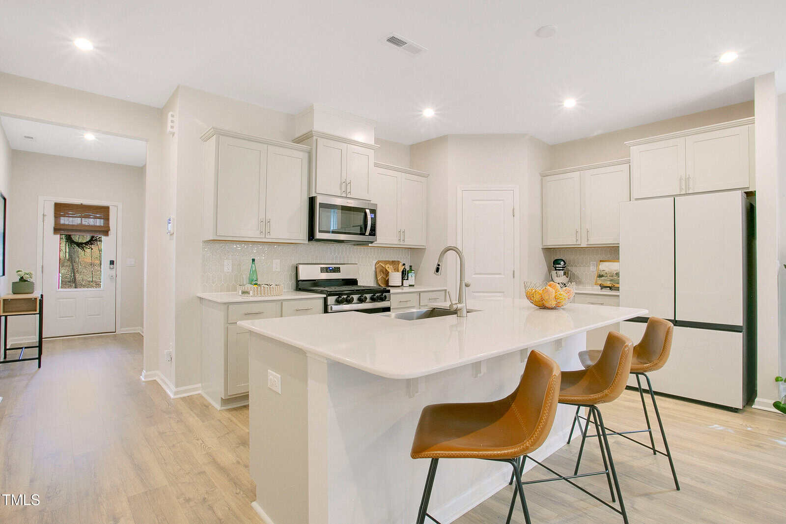 Modern kitchen with white cabinets, stainless steel appliances, a large island with a white countertop, and three brown barstools. Light wood flooring and neutral walls contribute to a bright, clean look.