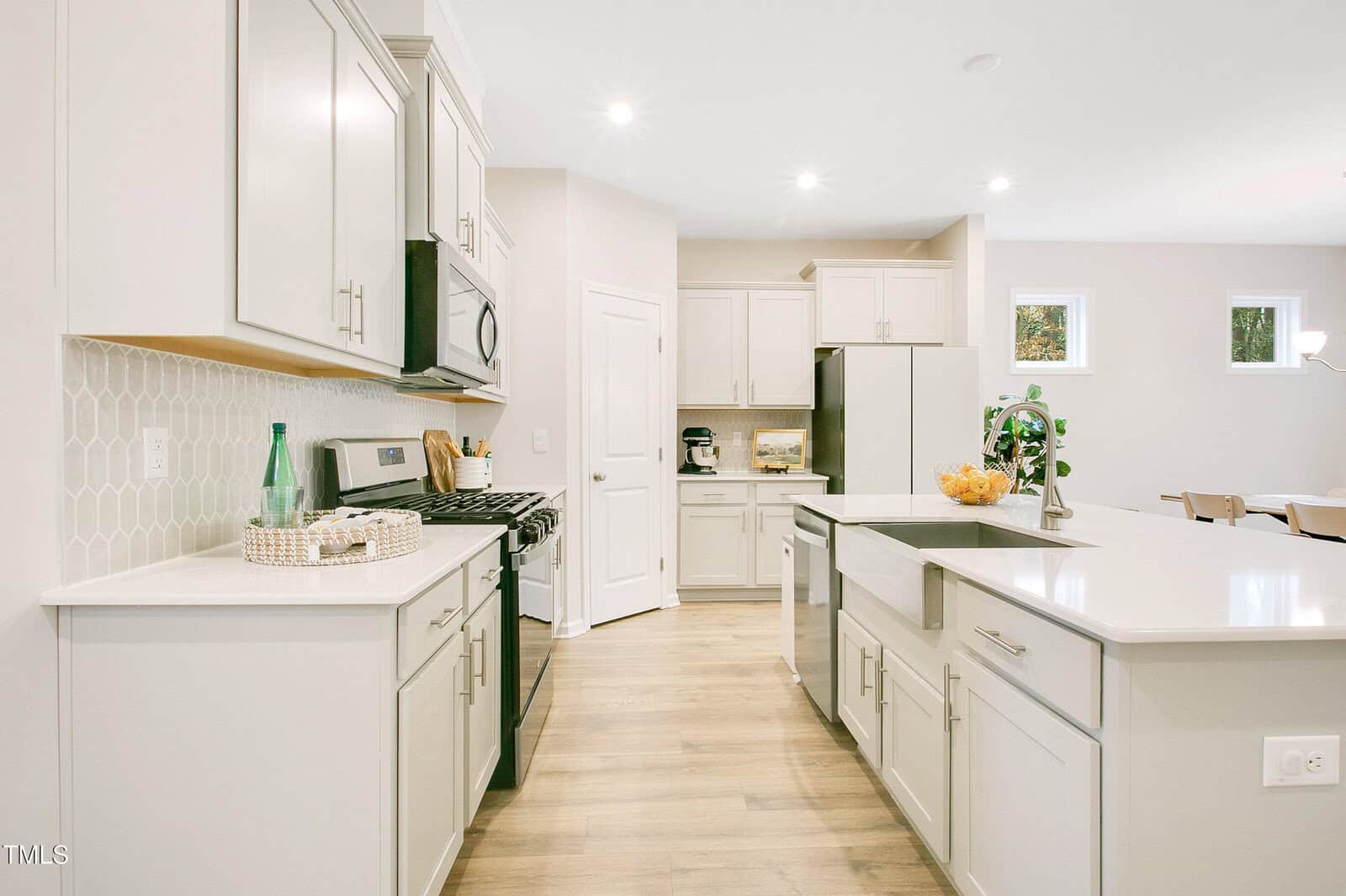 A modern kitchen with light-colored cabinets, stainless steel appliances, a farmhouse sink, a large island with seating, hexagonal tile backsplash, and wooden flooring. The space is well-lit with recessed lighting and natural light.
