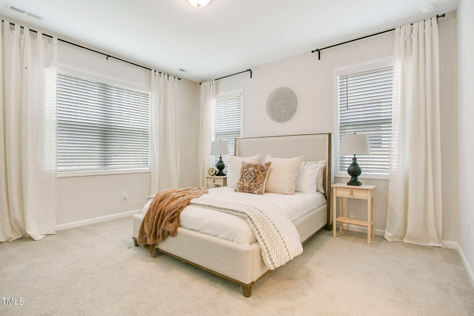 Bright bedroom with neutral tones, featuring a bed with white bedding, beige throw blanket, two nightstands with lamps, white curtains, and three windows with blinds. Walls and carpet are light-colored. Minimal decor.
