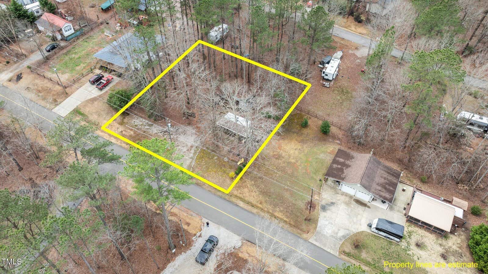 Aerial view of a wooded property outlined in yellow, located along a paved road in a residential area with nearby houses, driveways, vehicles, and several trees.