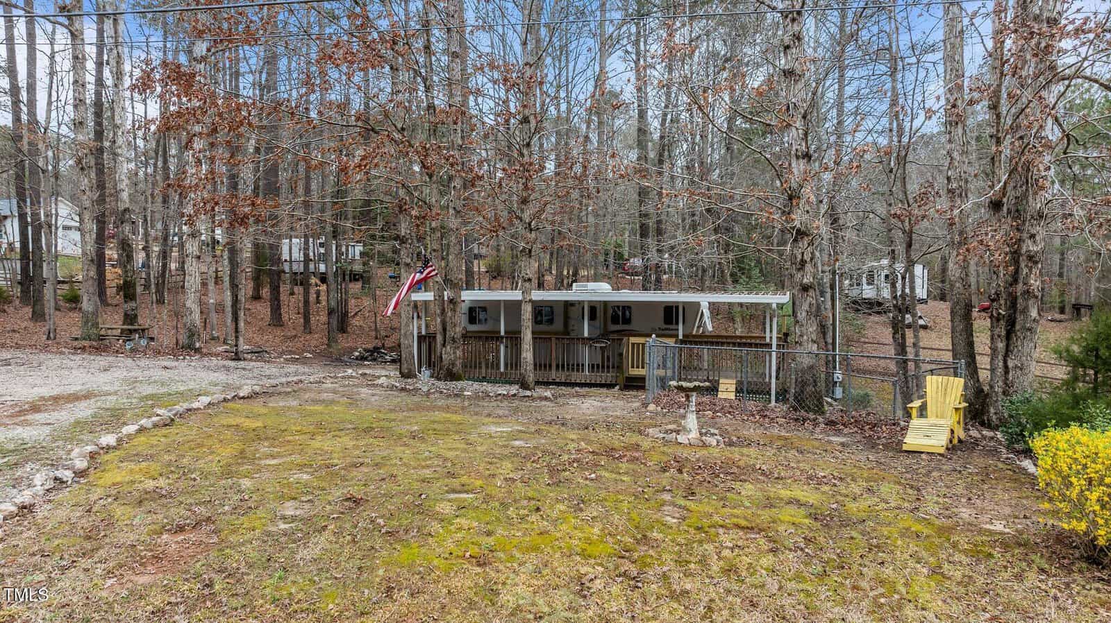 A small mobile home with a covered porch sits in a wooded area. The yard is mostly bare with patches of grass and moss. An American flag hangs by the porch, and two yellow chairs are placed near the right side.