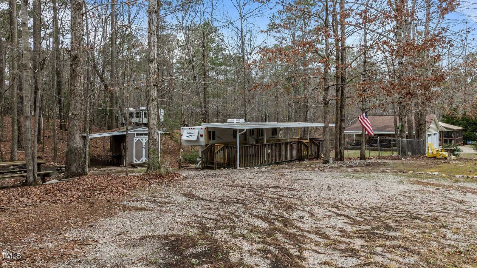 A trailer sits among trees on a gravel lot with a shed nearby. An American flag hangs near a small structure. Leaves cover the ground and a picnic table is visible on the left side of the image.