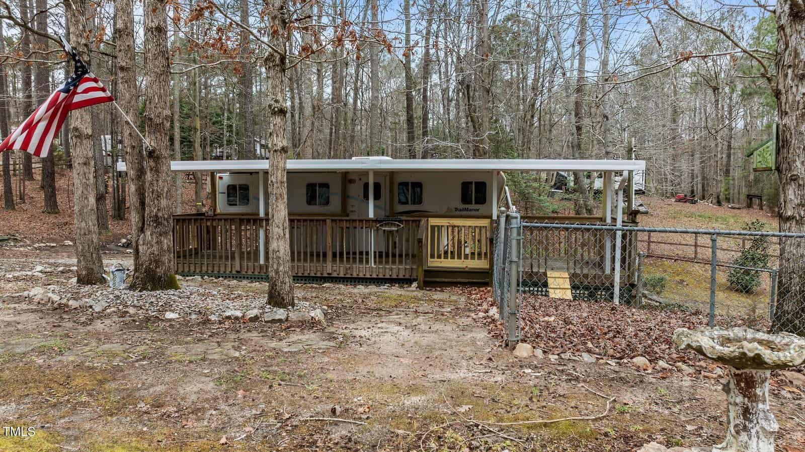 A white trailer with a wooden porch and railing is set in a wooded area behind a chain-link fence. An American flag hangs on the left, and leafless trees surround the trailer. Fallen leaves cover the ground.