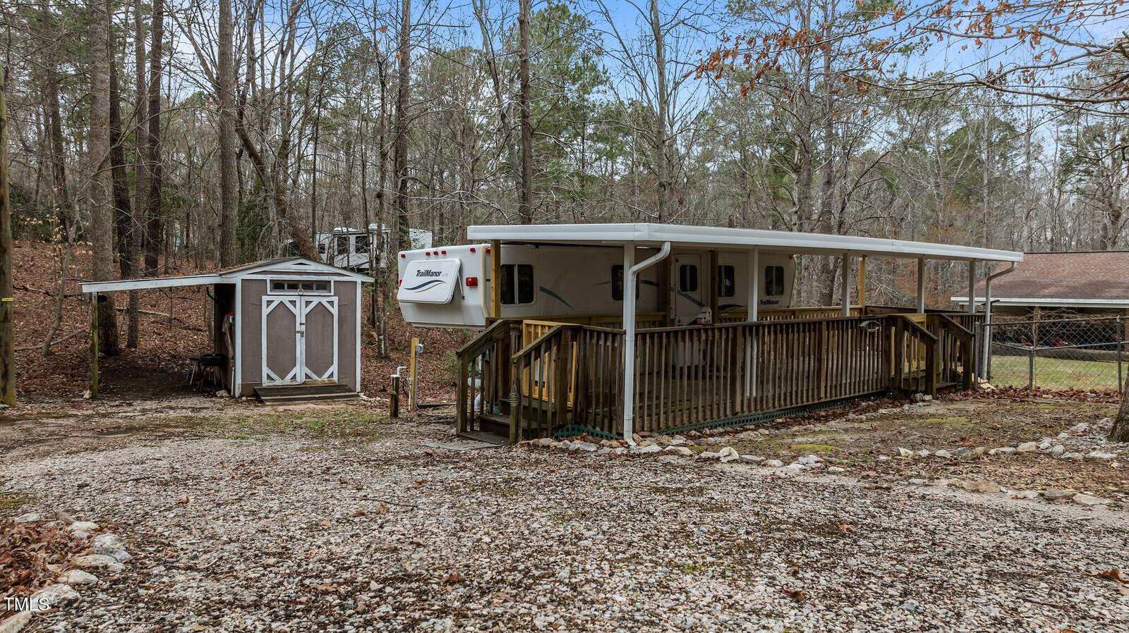 A white RV with an attached covered wooden porch is parked beside a small brown shed with white trim in a wooded, fenced area with leafless trees and a gravel driveway.