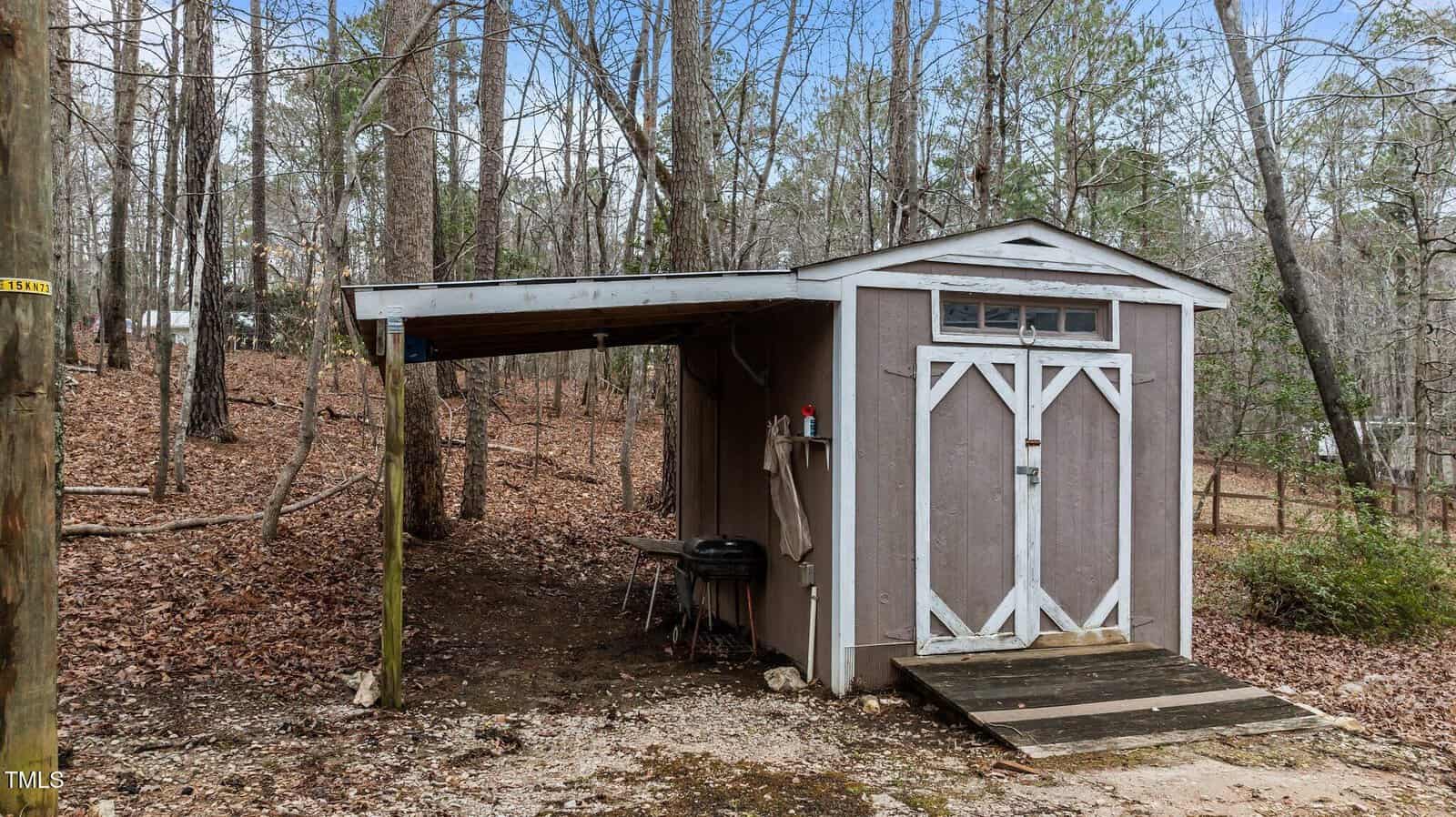 A small brown shed with white trim stands in a wooded area. It has double doors and an attached covered area with a grill and some items on a shelf. The ground is covered with leaves and there are trees in the background.