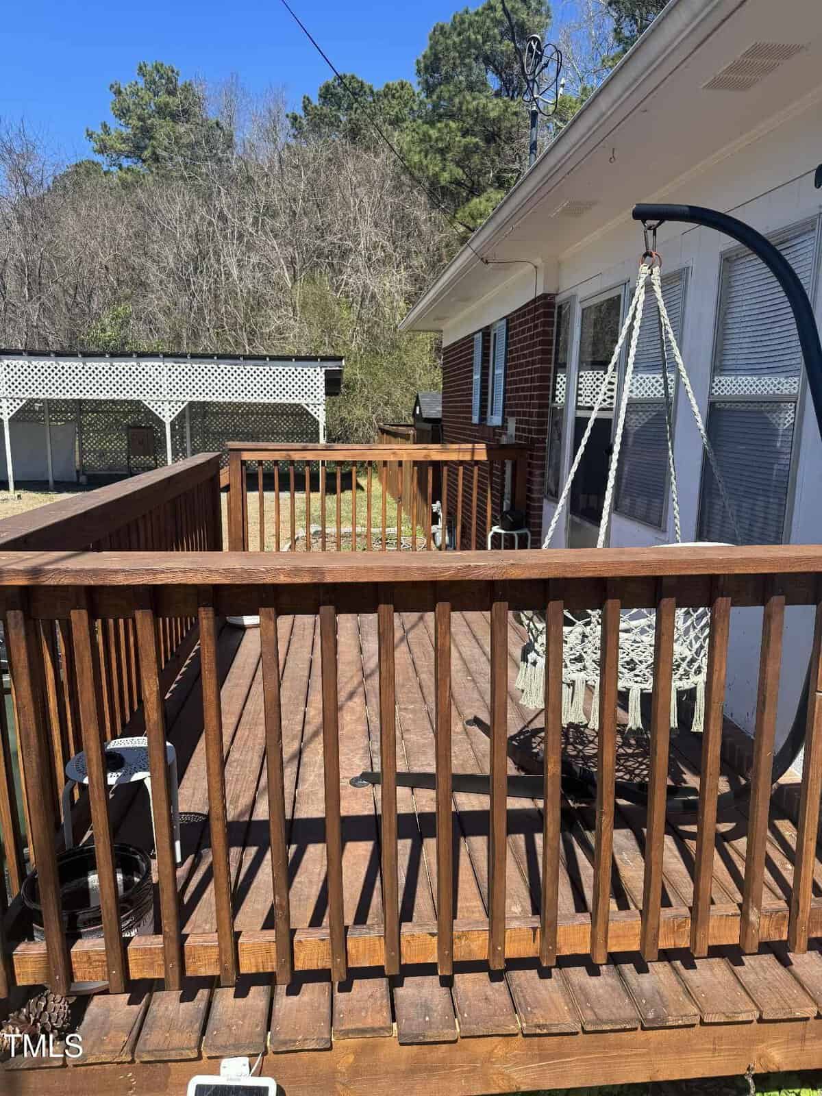 Wooden deck with railing attached to a white and brick house. The deck features a hanging swing chair. Trees and a lattice fence are visible in the background, along with a portion of a yard.