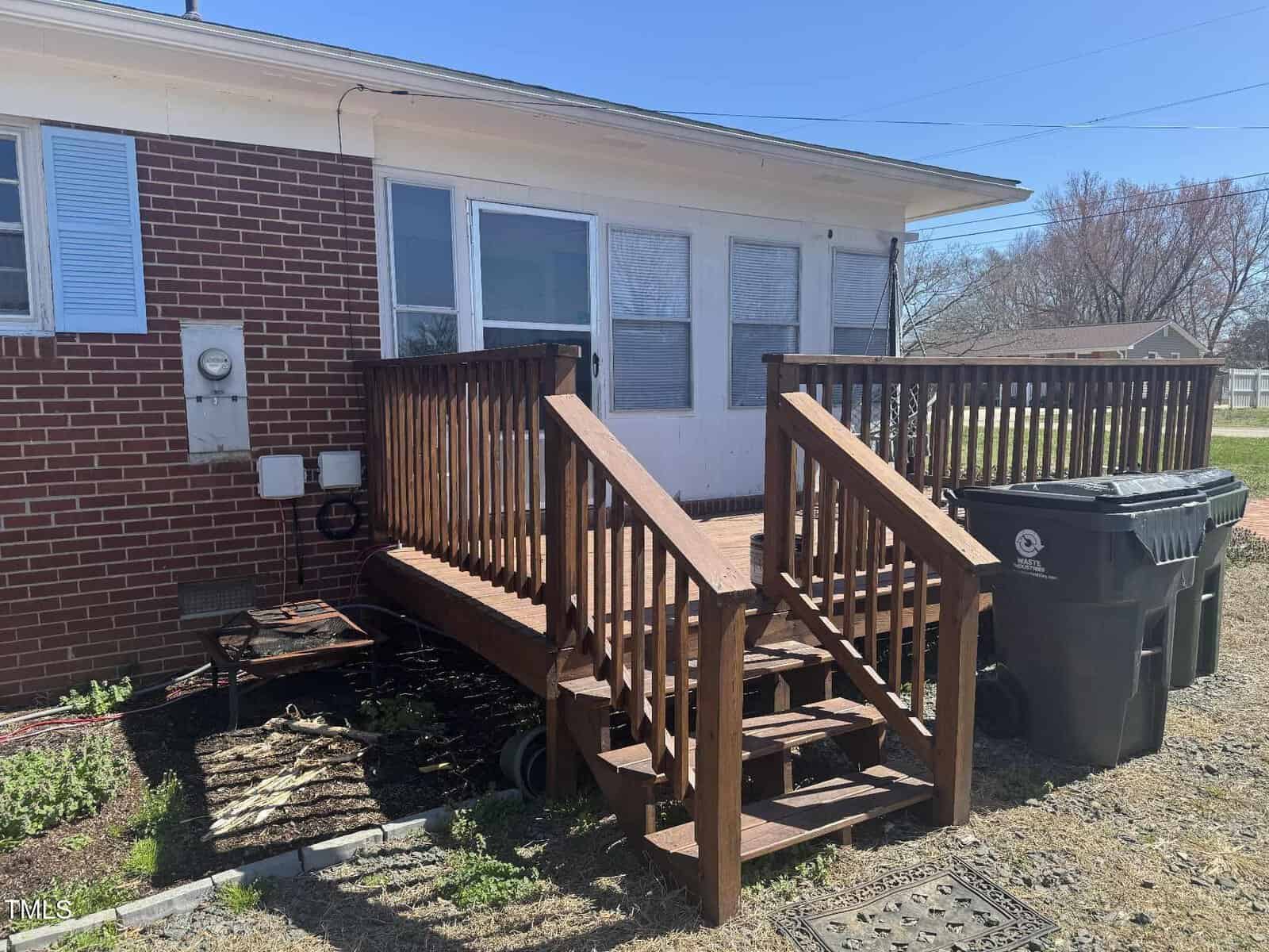 A small wooden deck with steps leads to a white door at the back of a brick house. Two large trash bins are positioned on the right side of the deck. The ground is a mix of dirt and grass, and a grill is placed on the left.