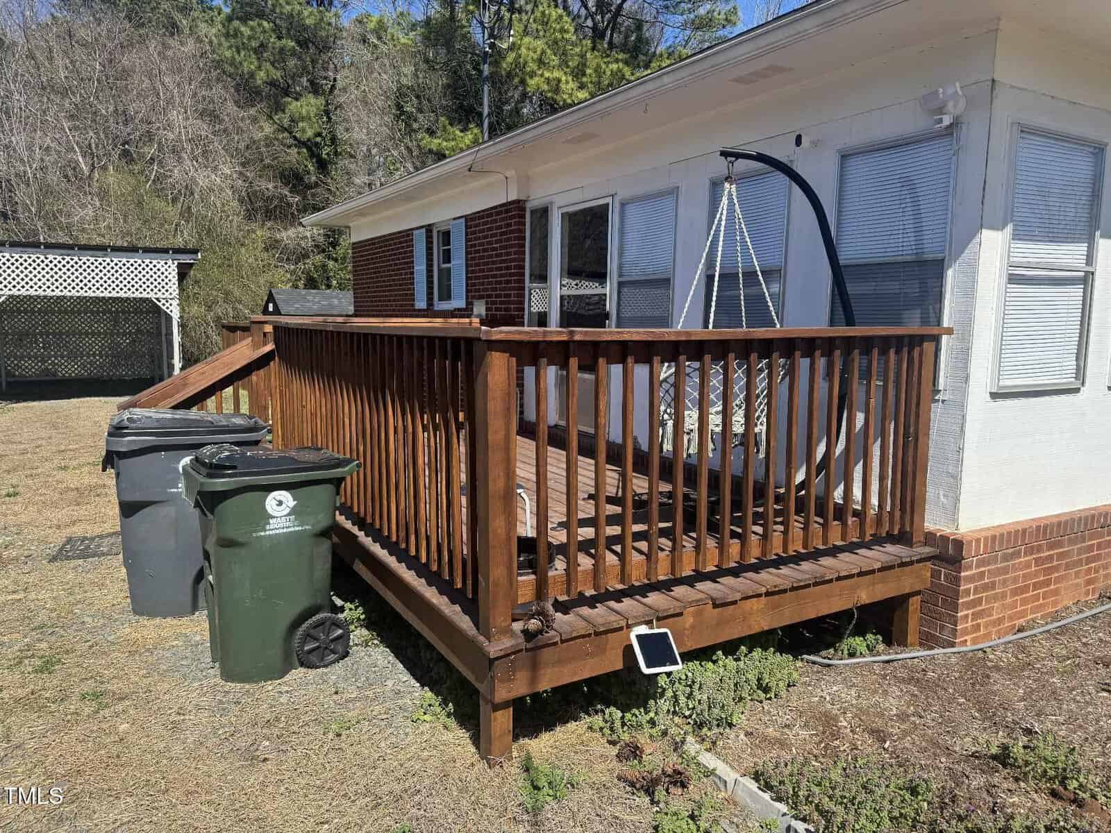 A small wooden deck with a railing attached to a house. A suspended chair hangs on the deck. Three trash bins are on the ground beside the deck. A sloping roof structure is visible in the background, surrounded by trees.