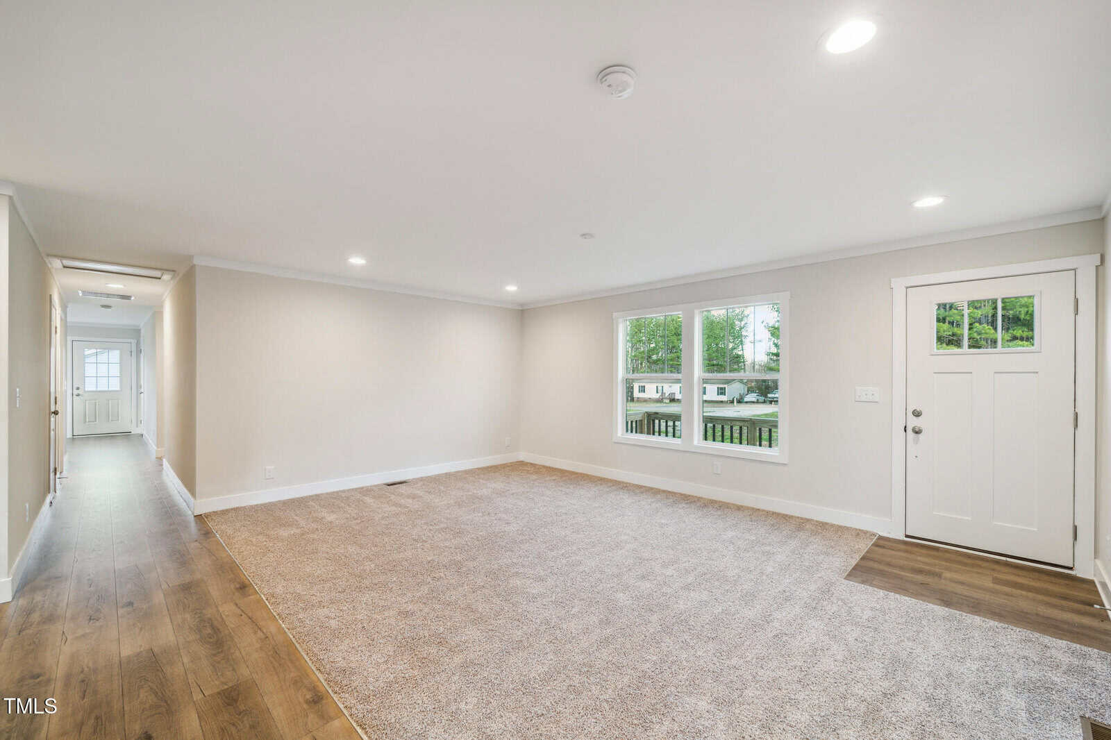 Spacious living room with light beige walls and a blend of carpet and wood flooring. A window and door are on the right, while a hallway leads to the back. Recessed lighting is installed in the ceiling.