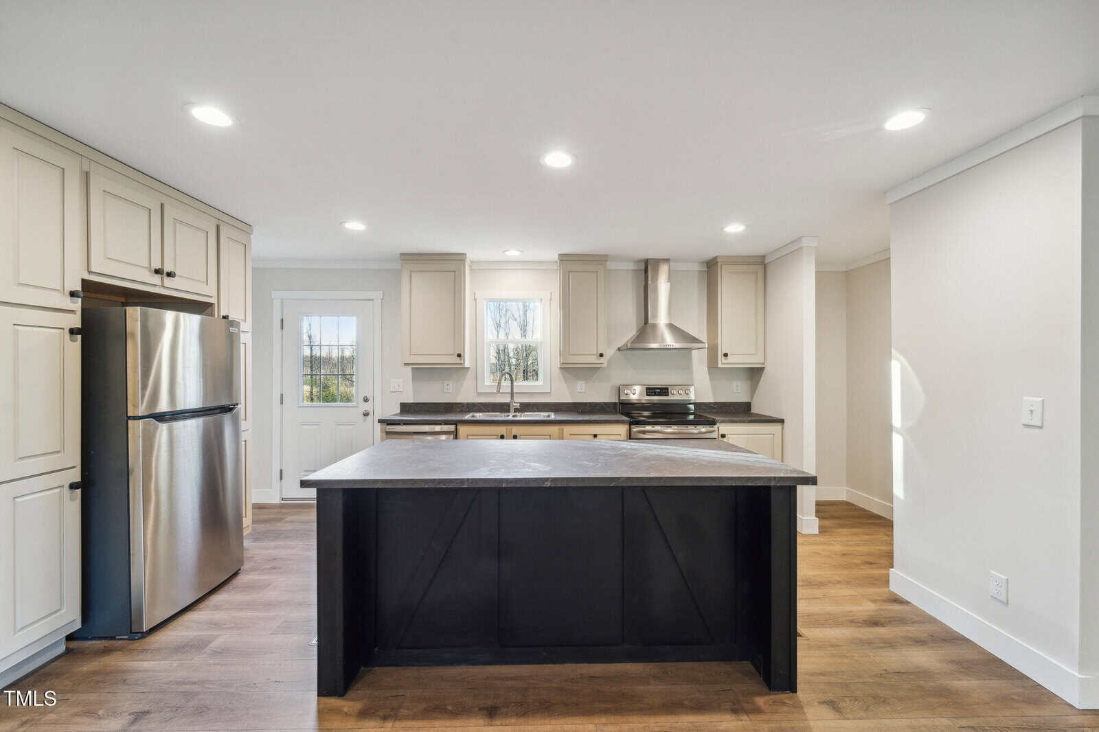 Modern kitchen with light-colored cabinets, stainless steel refrigerator, and oven. A large, dark island with a stone countertop is centered on a wooden floor. Recessed lighting is on the ceiling, and a door and windows bring in natural light.
