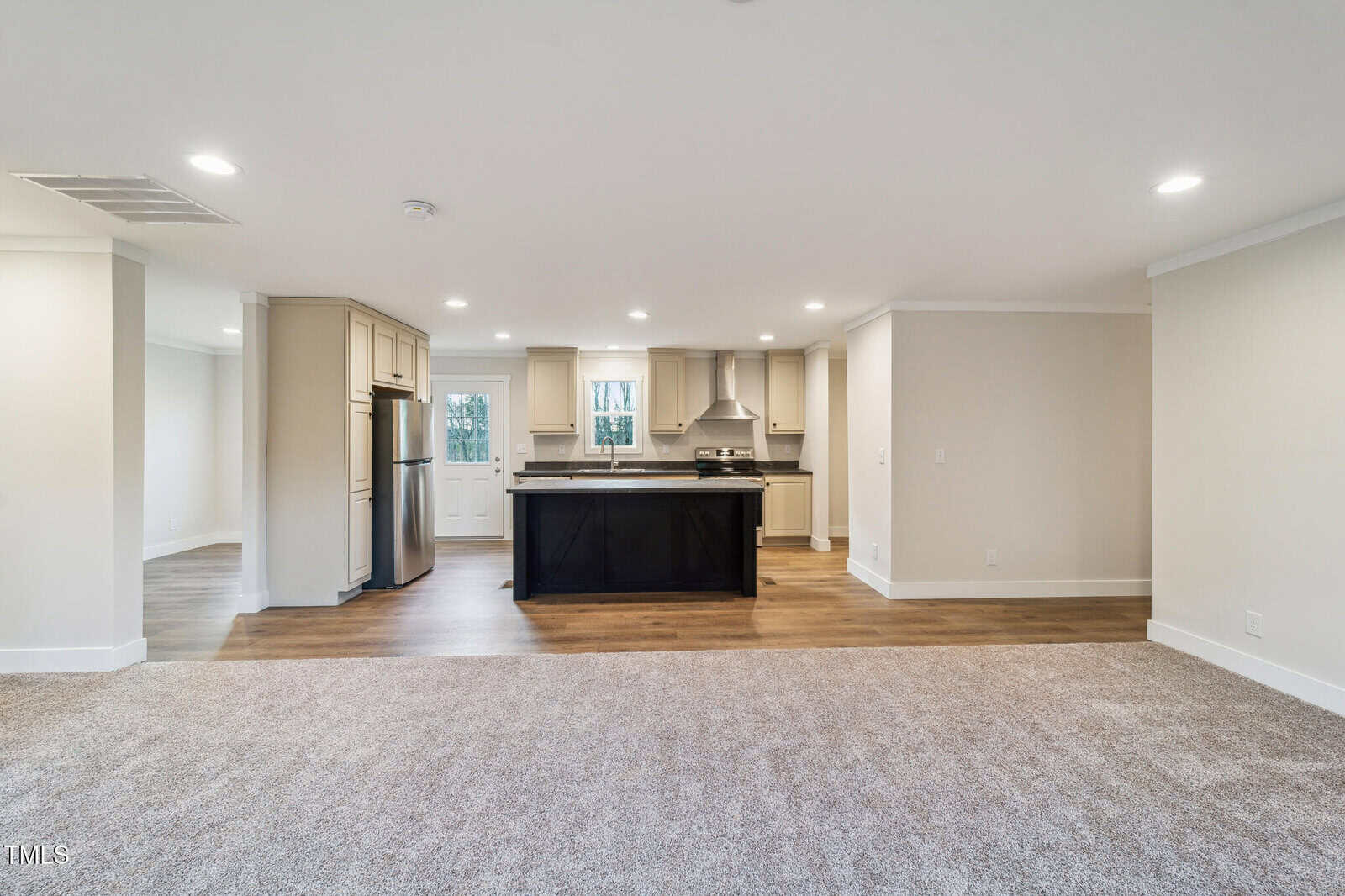 Open-concept living area featuring a spacious kitchen with black island, stainless steel appliances, and light wood cabinets. Carpeted area leads to a hardwood floor section. Neutral walls and recessed lighting provide a modern, airy feel.