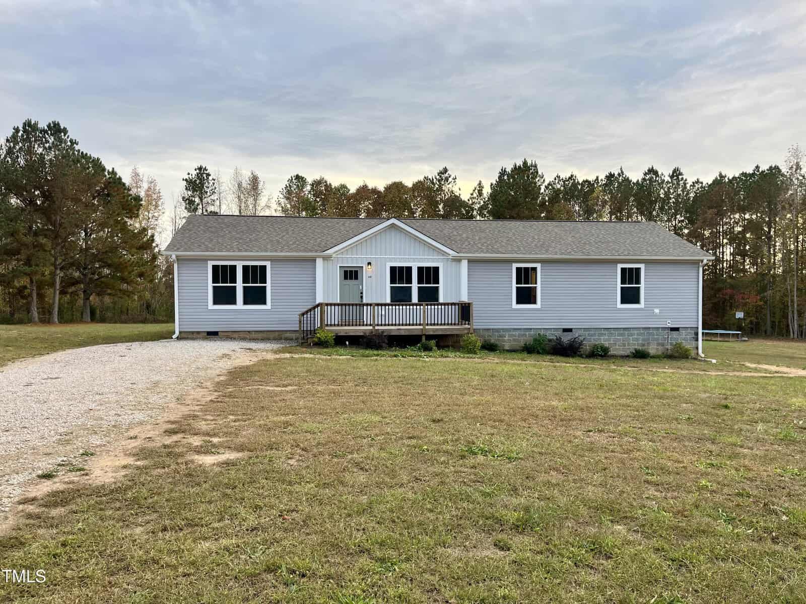 A single-story, gray house with white trim and a front porch sits on a spacious lawn at 349 Tristen Lane. A gravel driveway leads to the entrance of this charming Henderson residence. The background features a row of trees under a cloudy sky.